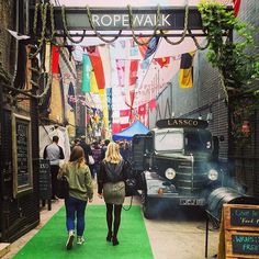 two women walking down the street in front of an old truck and flags hanging above them