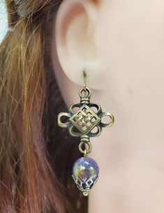 Stylized Celtic Knot Earrings in antique brass finish. Knots have domed center. A round purple and gold bead with beadcap dangles at bottom. Knots dangle from ear wires in antique brass finish. Designed and created by staff at Takeen Arts Studio. Item E1304. Antique Finish Brass Round Earrings, Antique Gold Adjustable Nickel-free Earrings, Bohemian Bronze Jewelry With French Hook, Unique Brass Beaded Earrings, Vintage Bronze Earrings With Round Beads, Bronze Earrings With Lever Back Ear Wires, Vintage Bronze Round Bead Earrings, Unique Brass Earrings With Round Beads, Bronze Lever Back Earrings