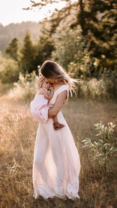 a woman in a white dress is holding a baby and looking at the ground with trees in the background