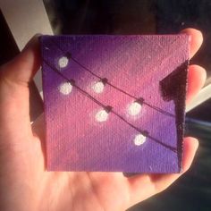 a hand holding up a small purple square with white dots on it and string lights in the background