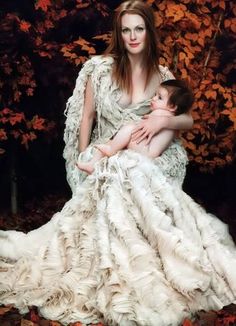 a woman holding a baby in a white dress with leaves on the ground behind her