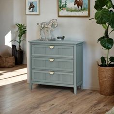 a blue dresser with three drawers in front of a painting on the wall next to a potted plant