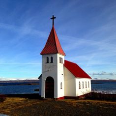 a white church with a red roof and cross on top by the water's edge