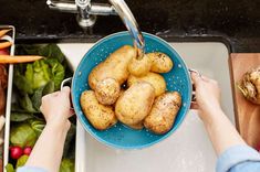 a person washing potatoes in a blue bowl on a kitchen sink next to fresh vegetables