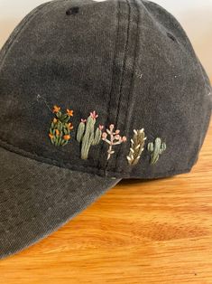 a baseball cap with embroidered cactus designs on the front and back side, sitting on a wooden surface