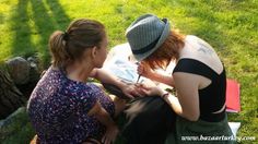 two women sitting on the grass with one writing something in her hand while the other writes