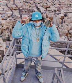 a man wearing a blue hat and jacket standing on top of a building with his hands in the air