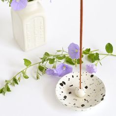 a white vase with purple flowers in it and a wooden stick sticking out of it