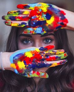 a woman with her hands painted in different colors and shapes, covering her eyes from the sun