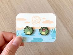 someone is holding up some cute little frog ear studs that are on a card