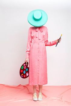 a woman in a pink dress and blue hat holding a small purse with her right hand