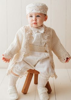 a baby sitting on top of a wooden chair wearing a white suit and bonnet hat