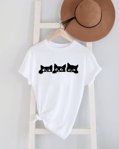 a white t - shirt with black cats on it next to a hat and ladder