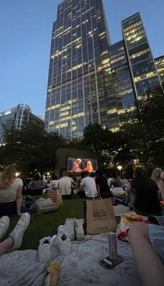 people are sitting on the grass in front of tall buildings at night, watching movies