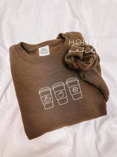 a brown t - shirt with coffee cups printed on the front and back, sitting on top of a white sheet