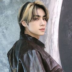 a young man with long hair wearing a leather jacket and looking at the camera while standing in front of a wall