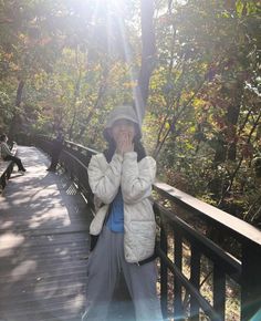 a woman covers her face while standing on a bridge in the woods with trees behind her