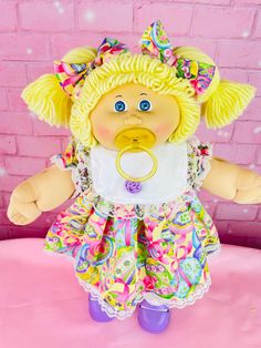 a baby doll with blonde hair and blue eyes on a pink surface in front of a brick wall