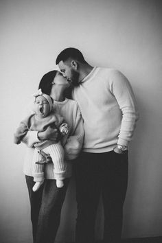 a man and woman kissing while holding a baby in front of a white wall with their hands on each other's heads