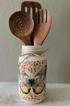 two wooden spoons are in a mason jar with butterflies and flowers painted on it