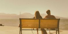 two people are sitting on a bench by the water with a dog looking at them