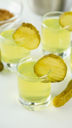 three glasses filled with yellow liquid next to a pickle on the side and other food items