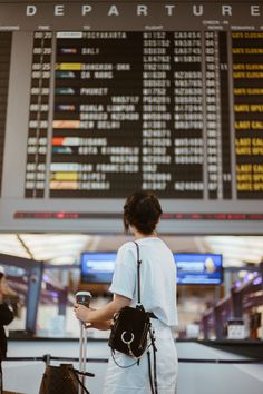 a woman standing at an airport with her luggage and looking at the departure board in the background