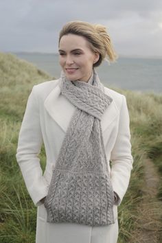 a woman standing in front of the ocean wearing a gray knitted scarf and white coat