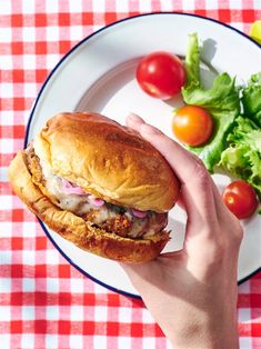 a hand holding a sandwich over a plate with salad and tomatoes on the tablecloth