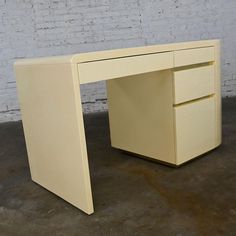 a white desk with two drawers in front of a brick wall and concrete flooring