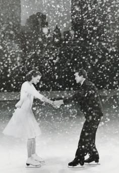 two people skating on an ice rink with snow falling all over the ground and buildings in the background