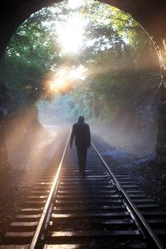 a man walking down train tracks towards the light at the end of the tunnel with trees in the background