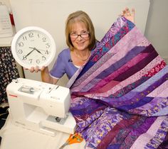 a woman is holding up a quilt with a sewing machine in front of her and a clock on the table