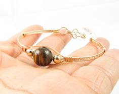 This beautiful bangle is handmade with a 10mm Tiger Eye Gemstone, two 14k gold filled beads and is wire wrapped with 14k gold filled wire. It is a fun bracelet and looks lovely on the wrist. To get the perfect fit, measure your wrist and add one half inch. Wire Wrapped Gold Bangle Bracelet As Gift, Gold Wire Wrapped Spiritual Bangle, Gift Wire Wrapped Round Bangle, Gift Round Bangle, Wire Wrapped Gold Bracelet As Gift, Gold Wire Wrapped Beaded Bracelets For Healing, Fun Bracelet, Brown Bracelet, Tiger Eye Bracelet