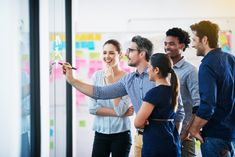 a group of people standing around each other in front of a whiteboard with sticky notes on it