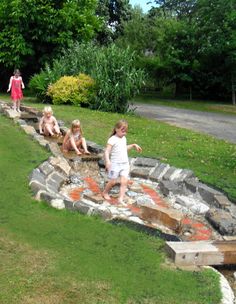 some kids are playing in the water near rocks and grass, while others walk by