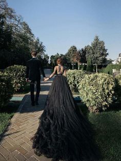 a man and woman in formal wear walking down a path holding hands, dressed in black