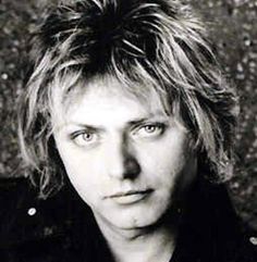 a black and white photo of a man with blonde hair looking at the camera, in front of a grungy wall