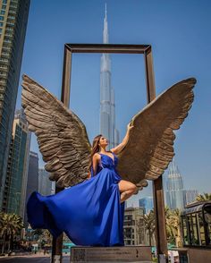 a woman in a blue dress is posing next to an angel statue with skyscrapers in the background