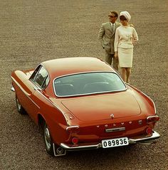 two people standing next to an old red car in the middle of a dirt field