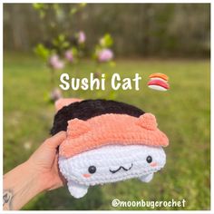 a hand holding up a small sushi cat plush toy in front of a green field