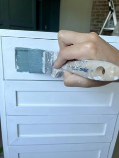 a person is holding a paintbrush and painting on a white cabinet with drawers in the background