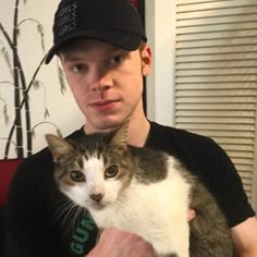 a man holding a cat in his arms and looking at the camera while wearing a hat