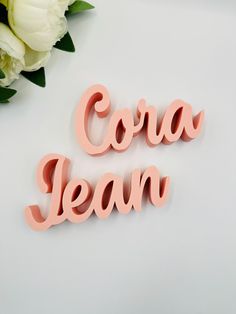 the word cora jean is made out of wood and sits next to some white flowers