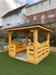 a small wooden shelter in the middle of a yard with grass and buildings behind it