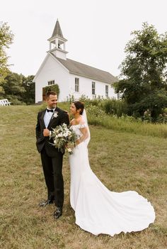 a bride and groom standing in front of a white church on their wedding day, looking at each other