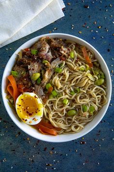 a white bowl filled with noodles, meat and veggies next to an egg