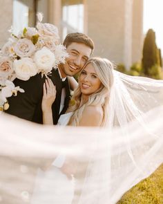 a bride and groom pose for a photo in front of their wedding venue with the veil blowing in the wind