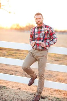 a man leaning against a white fence wearing a plaid shirt and bow tie with suspenders