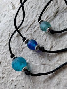 two necklaces with blue and green glass beads on black cord hanging from a stone wall